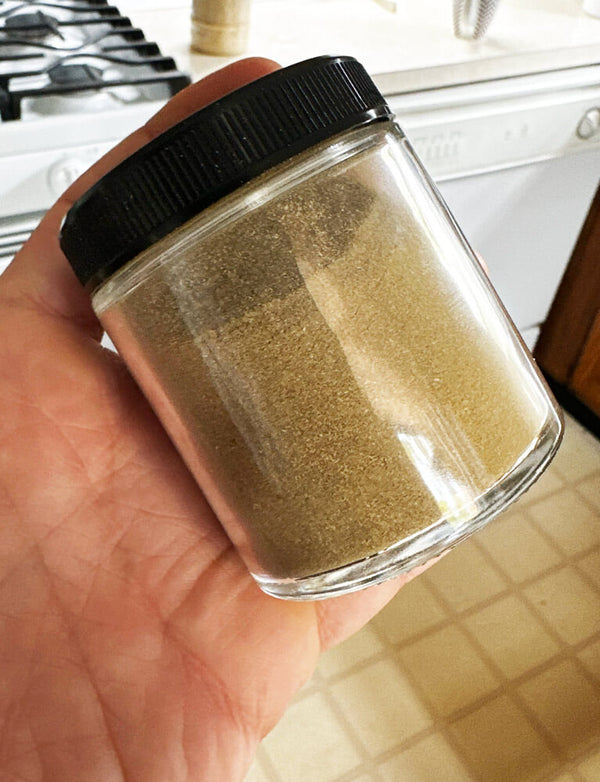 Hand holding a small glass jar with black lid containing a brown powder, kitchen counter in the background